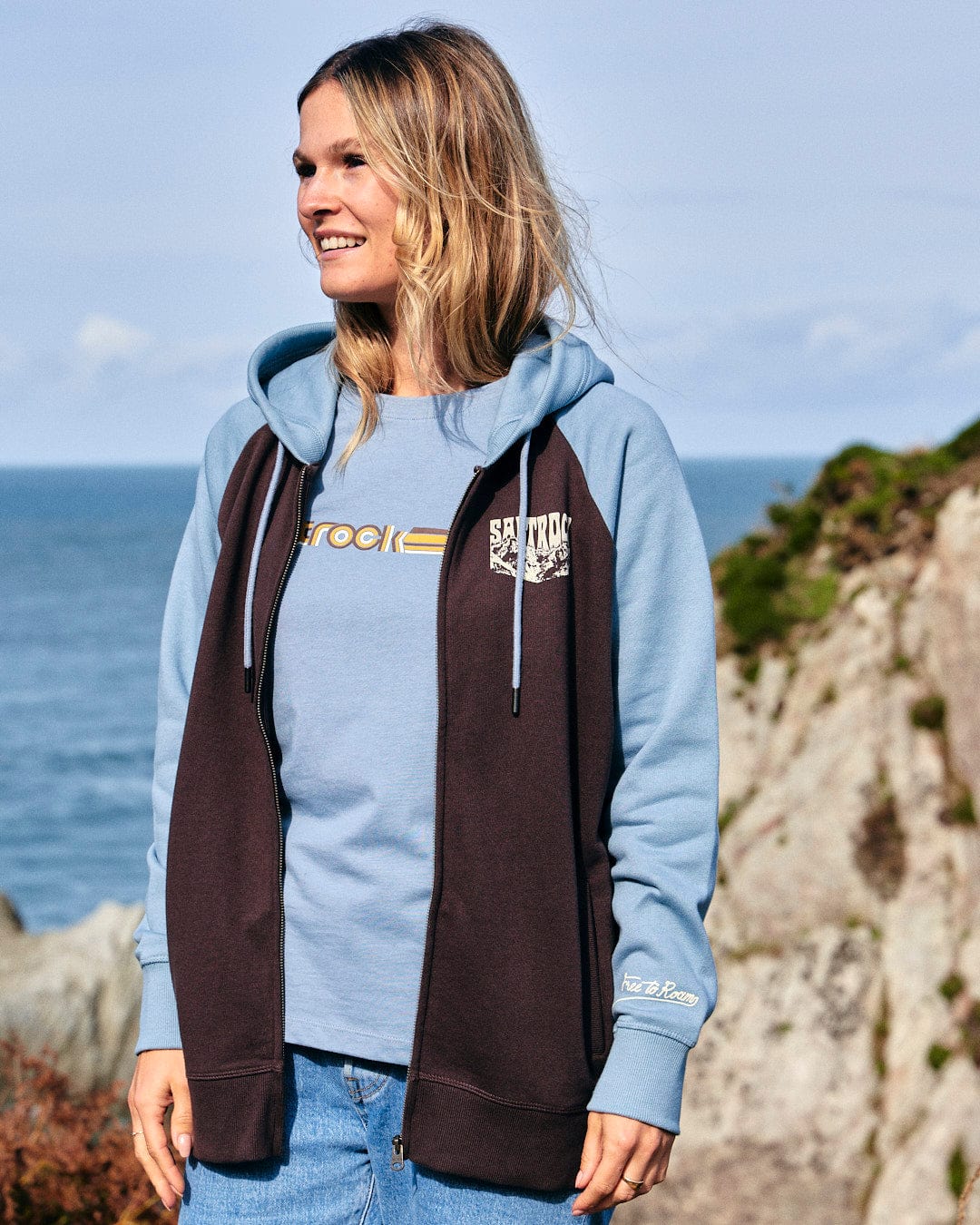 An adventurous woman wearing a blue and brown Saltrock Mountain Roaming - Womens Raglan Zip Hoodie - Brown standing next to the ocean.