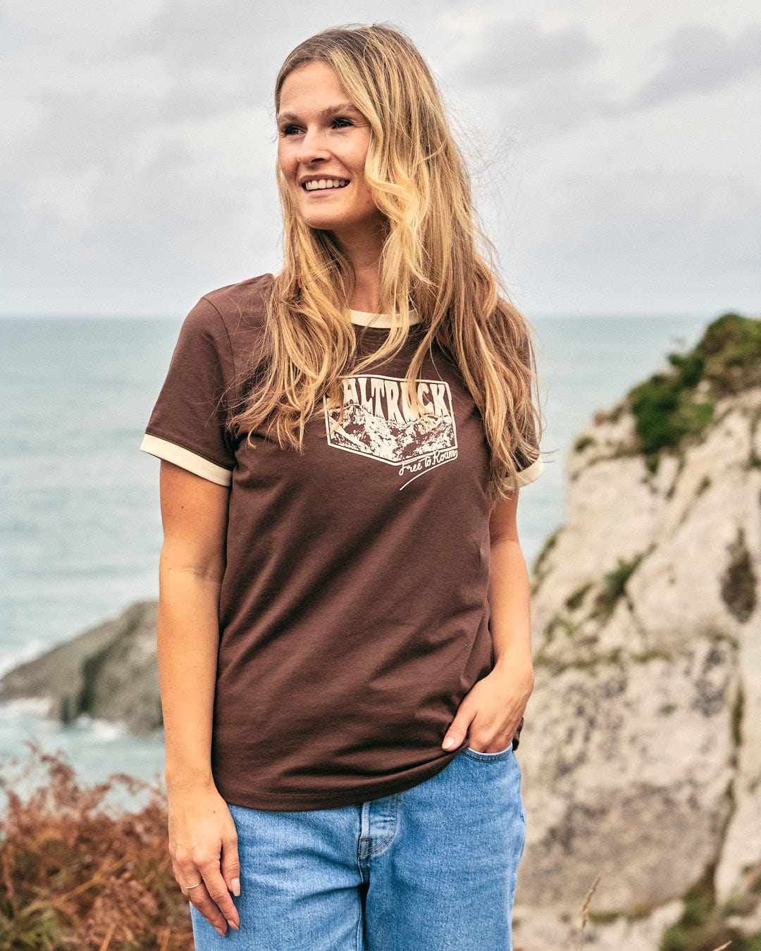 A woman wearing a Saltrock branded Mountain Roaming - Womens Short Sleeve T-Shirt - Brown standing by the ocean, exuding comfort.