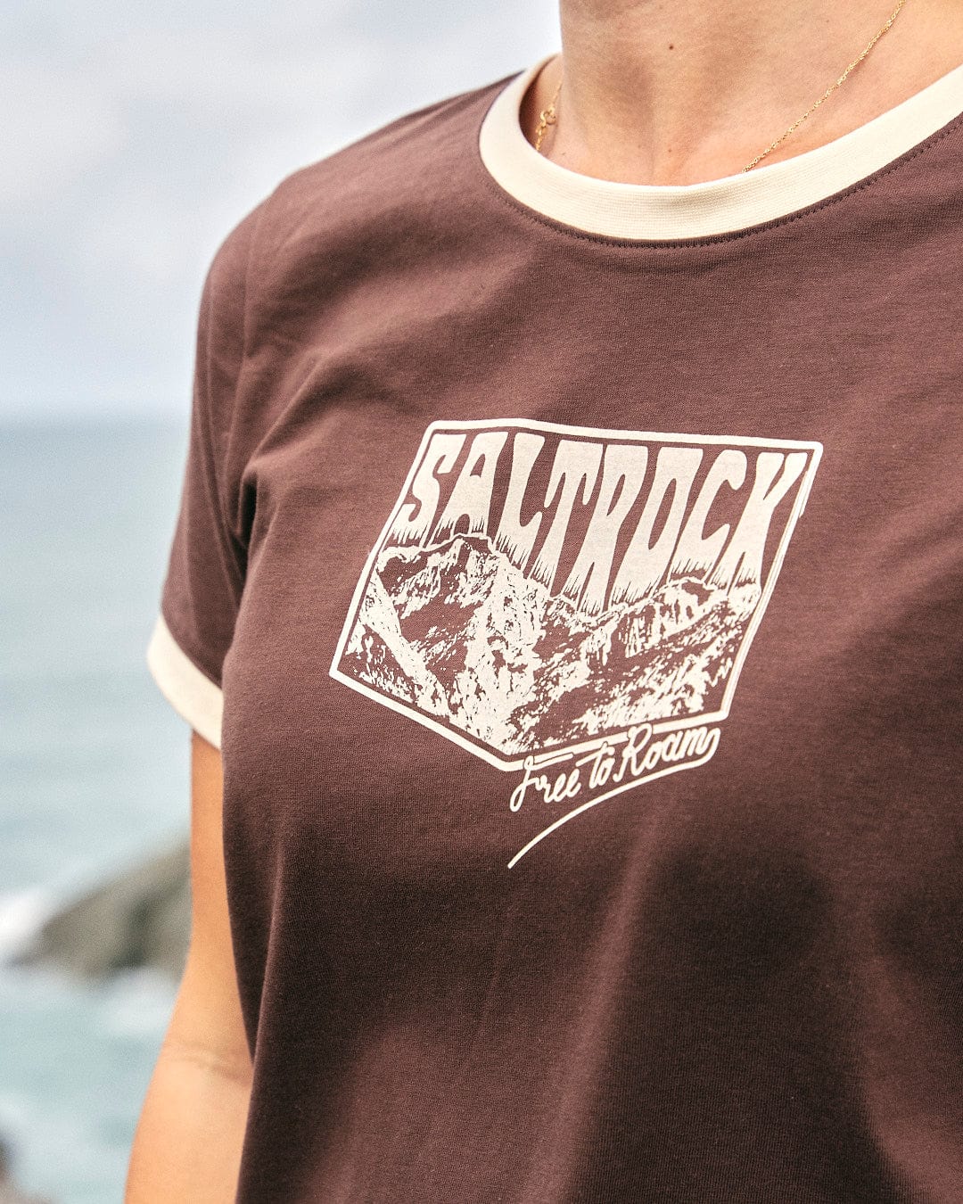 A woman standing in front of the ocean wearing a comfortable Mountain Roaming - Womens Short Sleeve T-Shirt - Brown adorned with Saltrock branding.