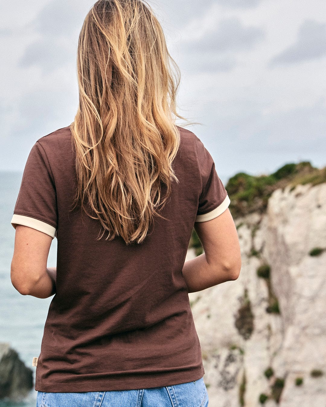 A woman wearing a Saltrock Mountain Roaming - Womens Short Sleeve T-Shirt - Brown, comfortingly looking at the ocean.