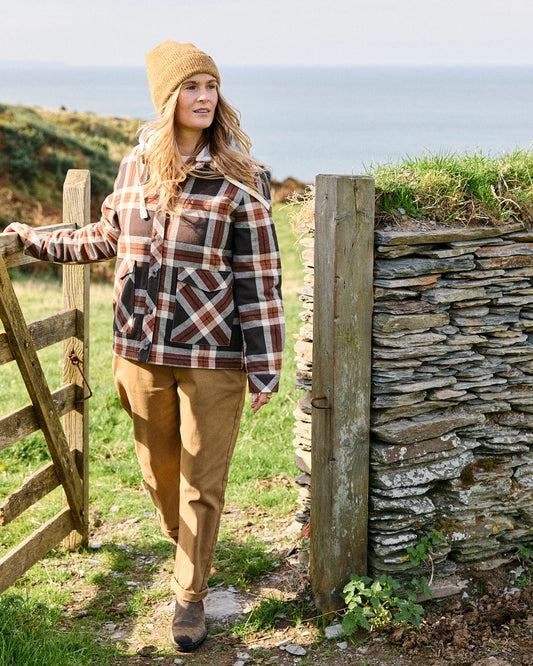 A person wearing the Saltrock Myla Women's Hooded Shacket in brown stands by a wooden gate near a stone wall, with grassy hills and the sea in the background.