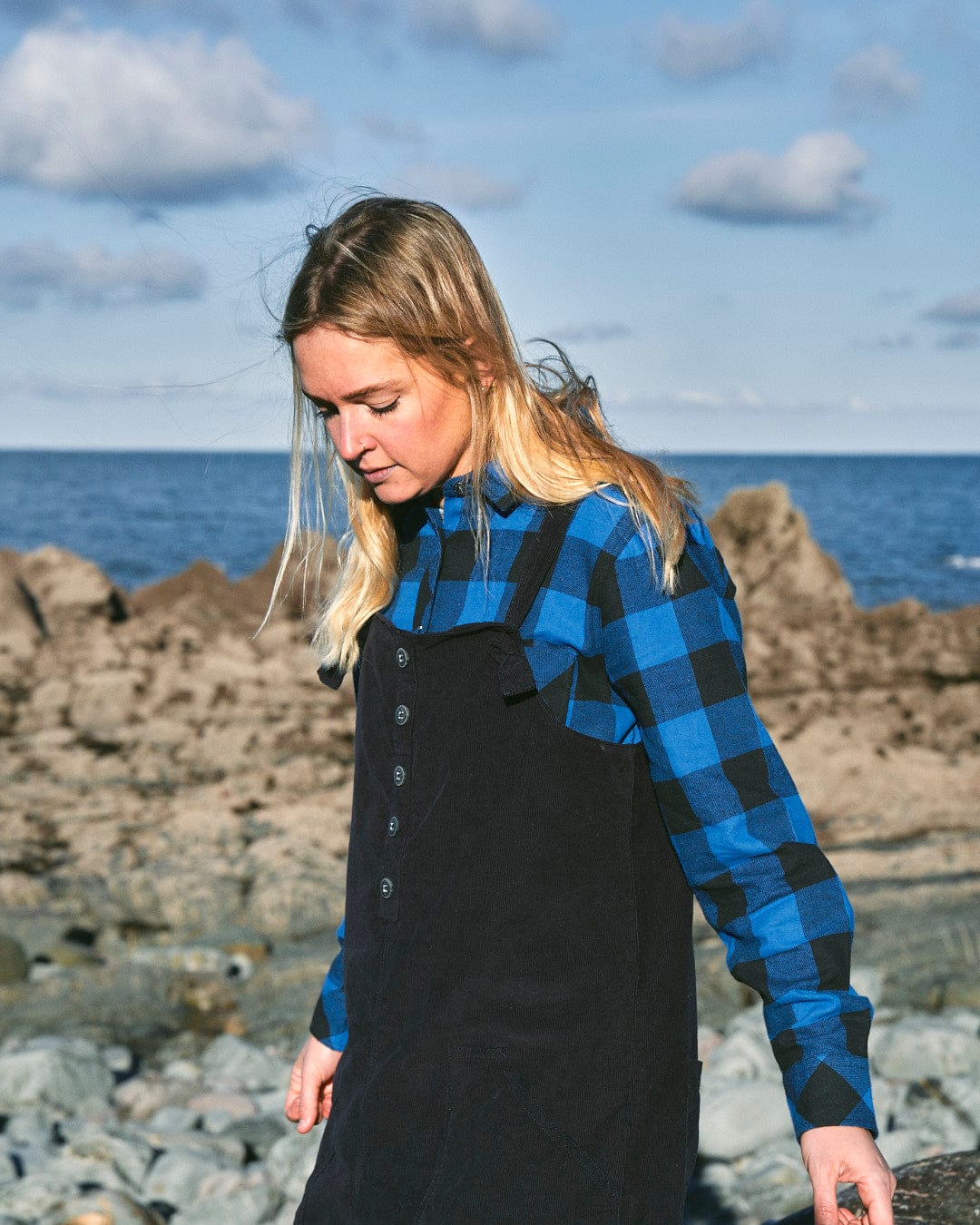 A woman wearing a stylish Saltrock Nancy - Womens Cord Dungaree - Dark Blue in a blue and black plaid shirt standing on rocks near the ocean.