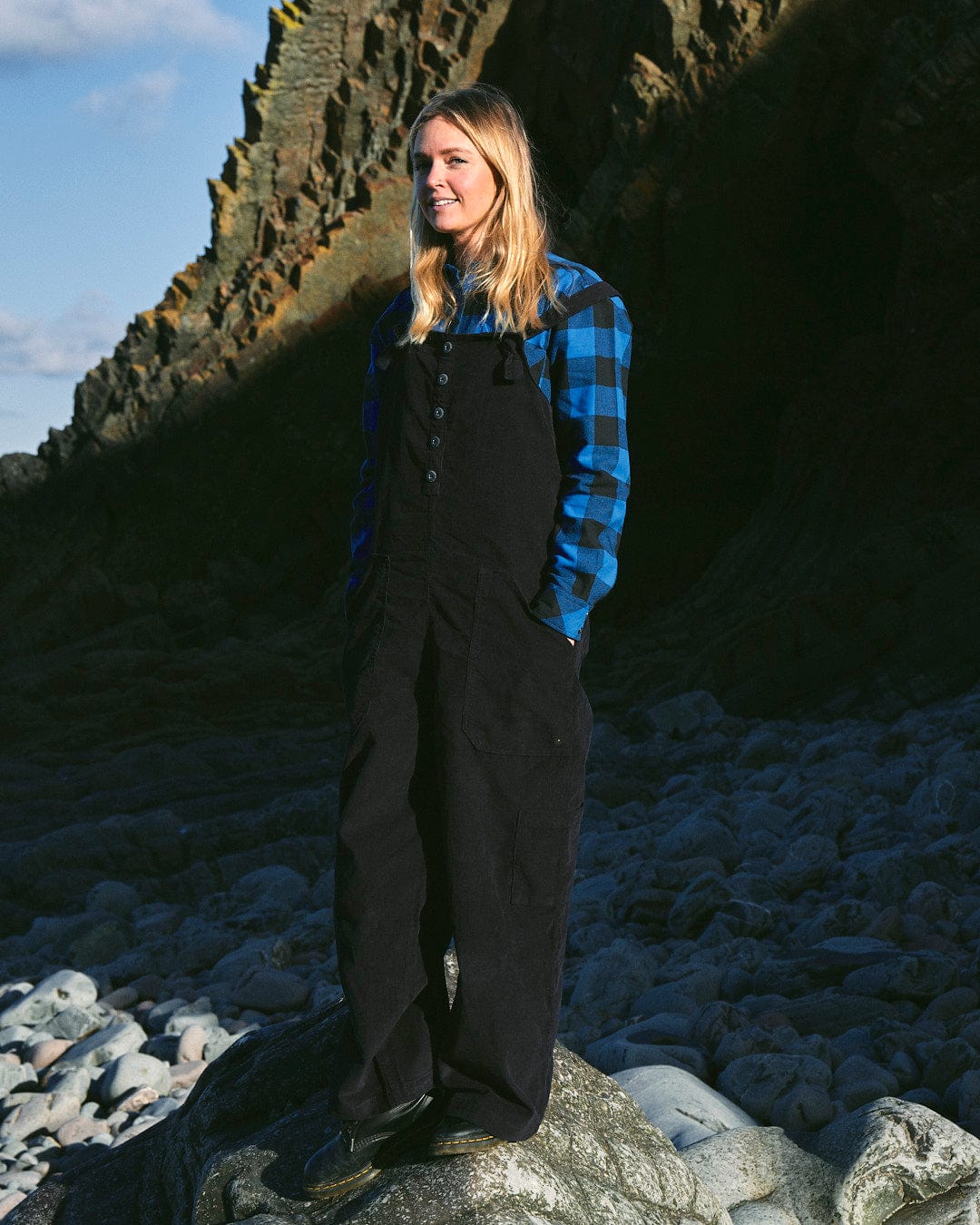 A fashion-conscious woman in Nancy - Womens Cord Dungaree - Dark Blue dungarees standing on rocks by Saltrock.