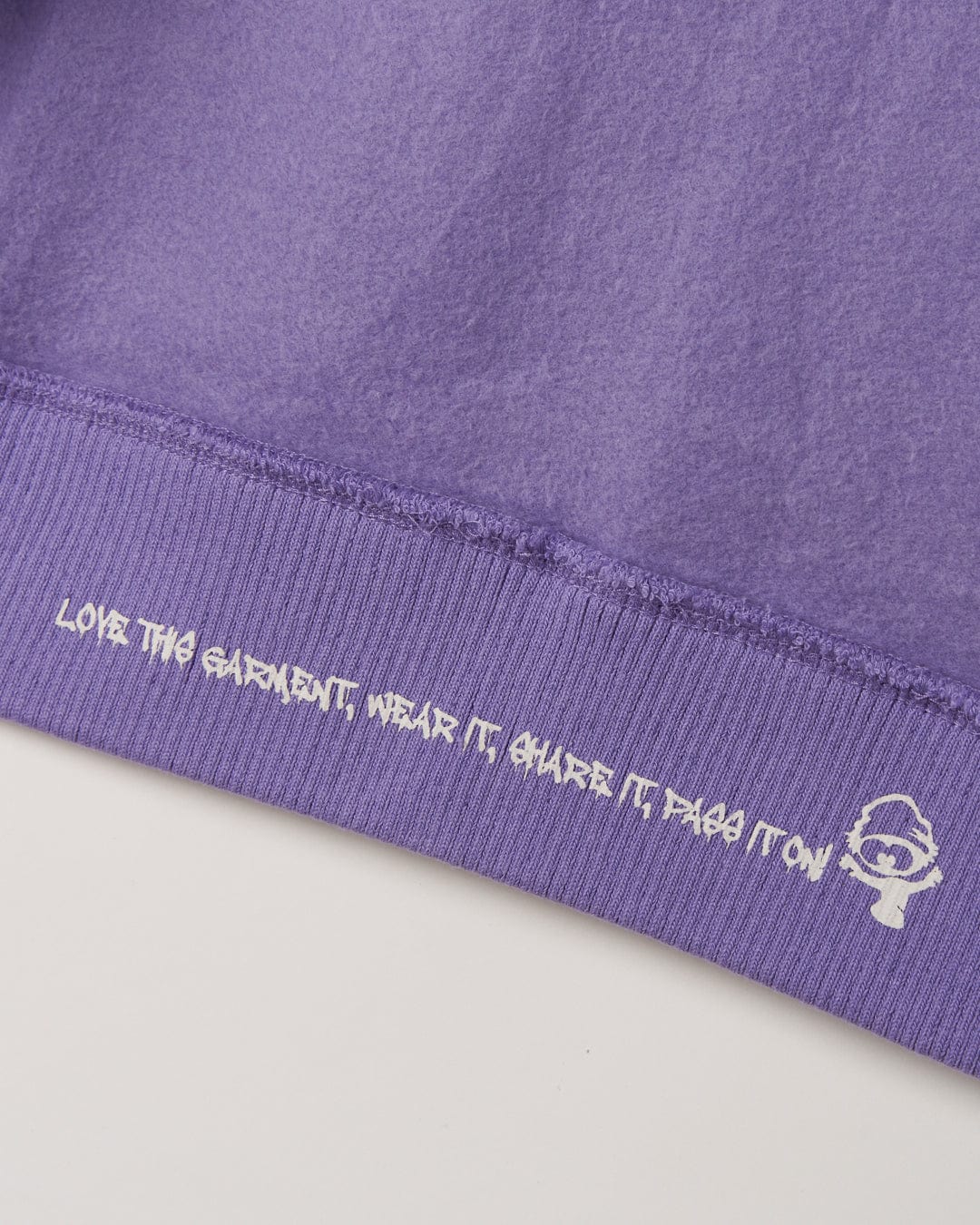 Close-up of a purple fabric with a slogan "love the garment. wear it. share it. pass it on" embroidered along the seam, accompanied by a small white Saltrock branded logo on the No Road No Problem - Recycled Kids Zip Hoodie - Purple.