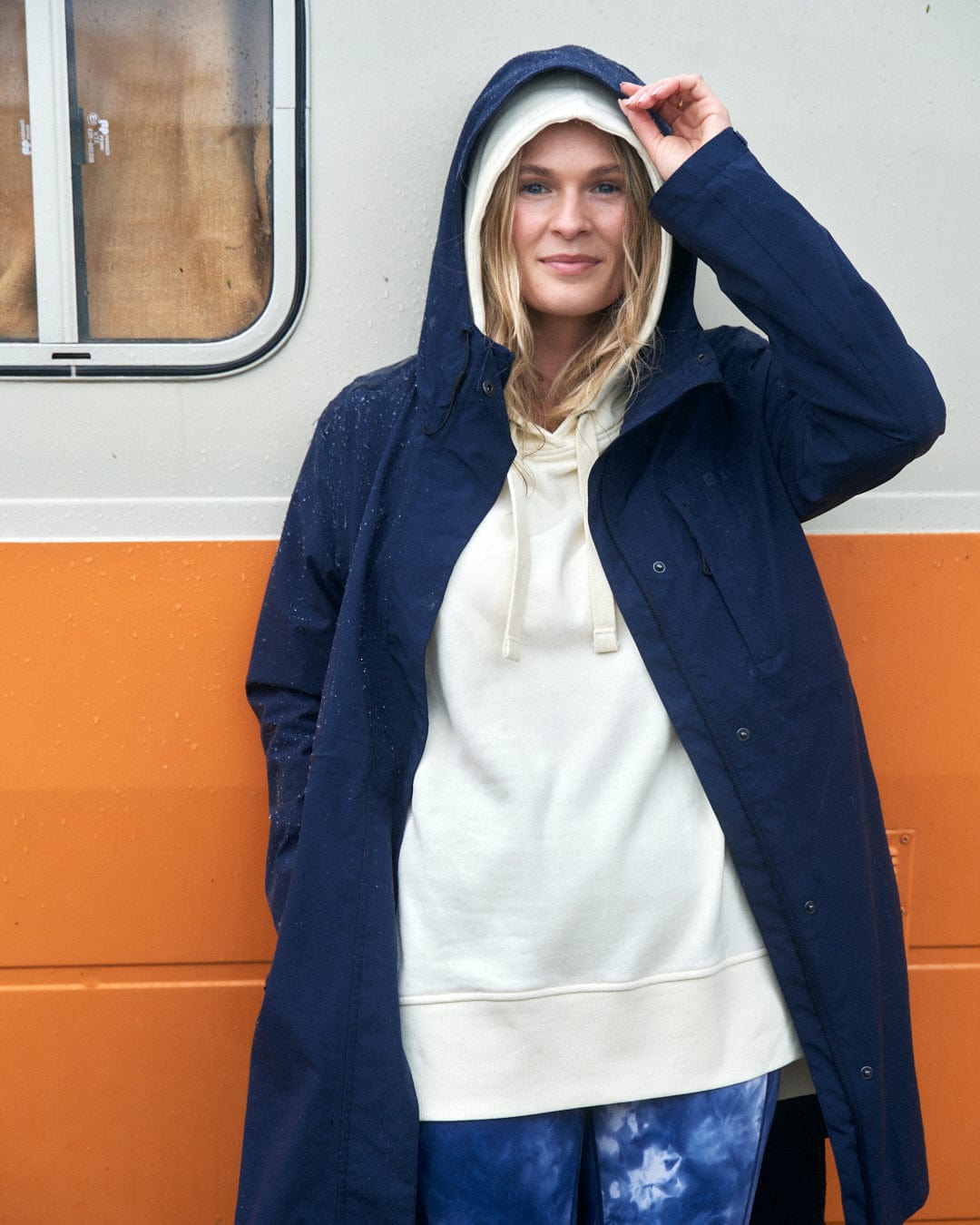 A smiling woman in a North West - Womens Waterproof Jacket - Blue by Saltrock, with an adjustable hood and hoodie, standing in front of an orange and white tram.