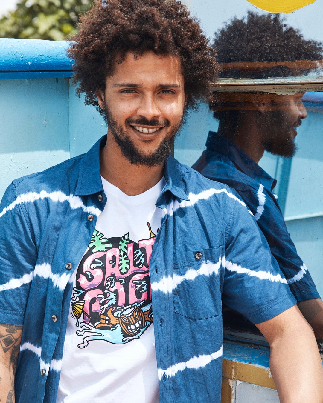 A young man with curly hair smiling at the camera, wearing a Saltrock Ocean - Mens Tie Dye Shirt in Blue and blue open shirt, leaning against a colorful wooden structure.