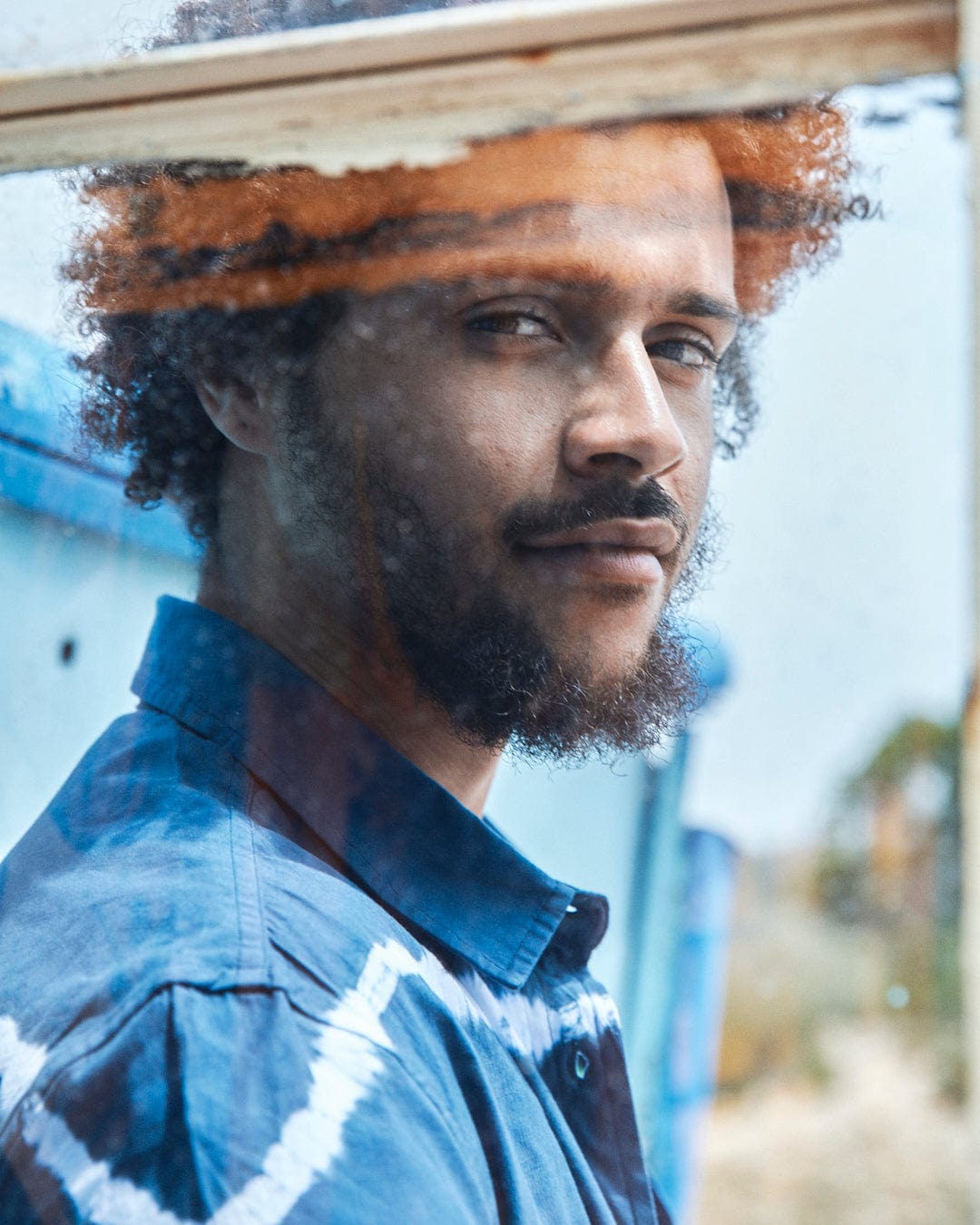 Man with curly hair looking through a glass window, wearing a Saltrock Ocean - Mens Tie Dye Shirt in Blue. Reflection visible on the glass.