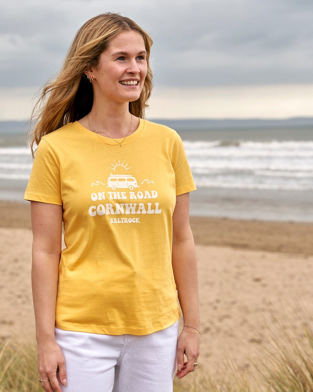A woman wearing a Saltrock On The Road Cornwall - Womens Short Sleeve T-Shirt - Yellow standing on the beach.