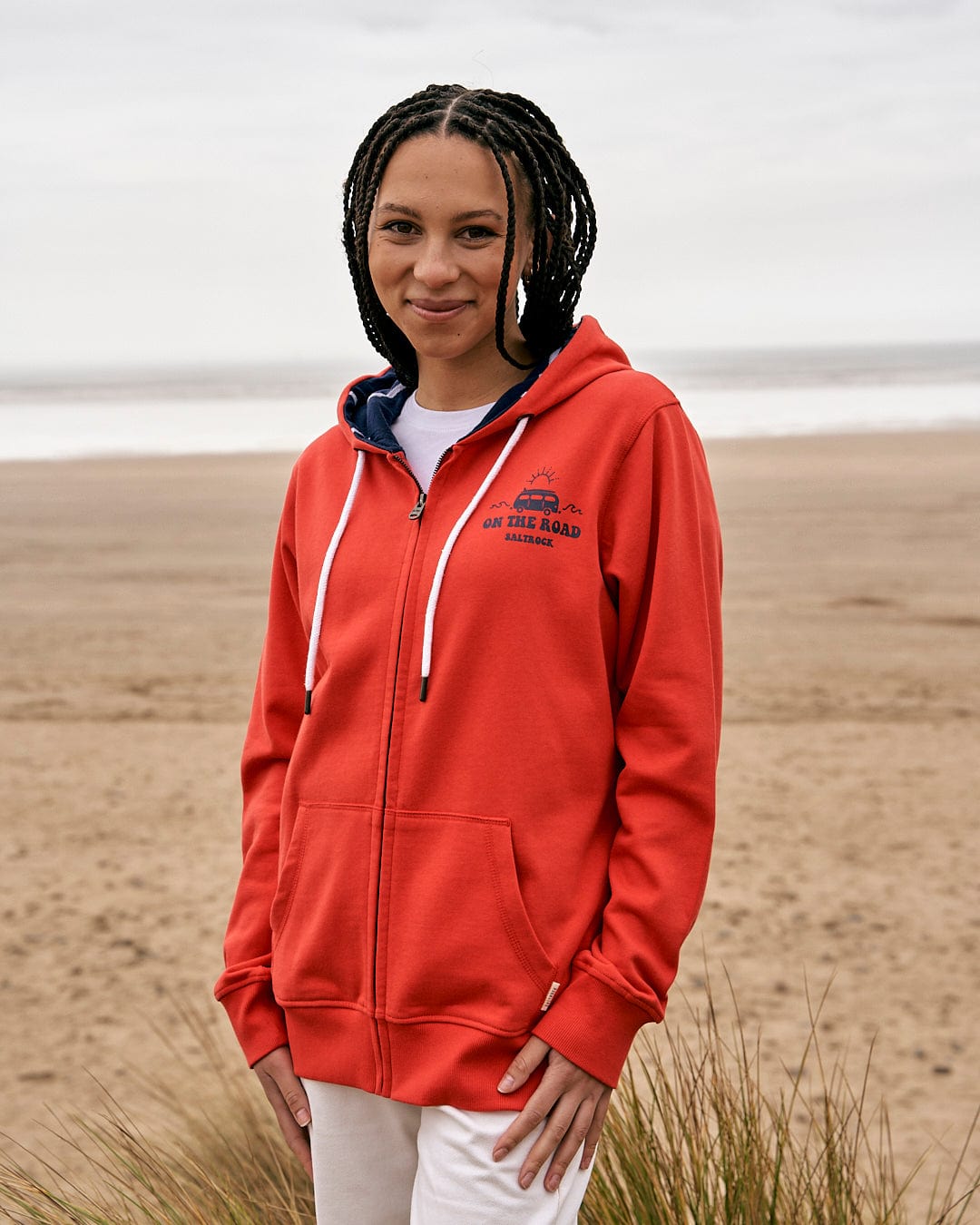 A woman in a Saltrock On The Road Devon - Womens Zip Hoodie - Red standing on a beach.