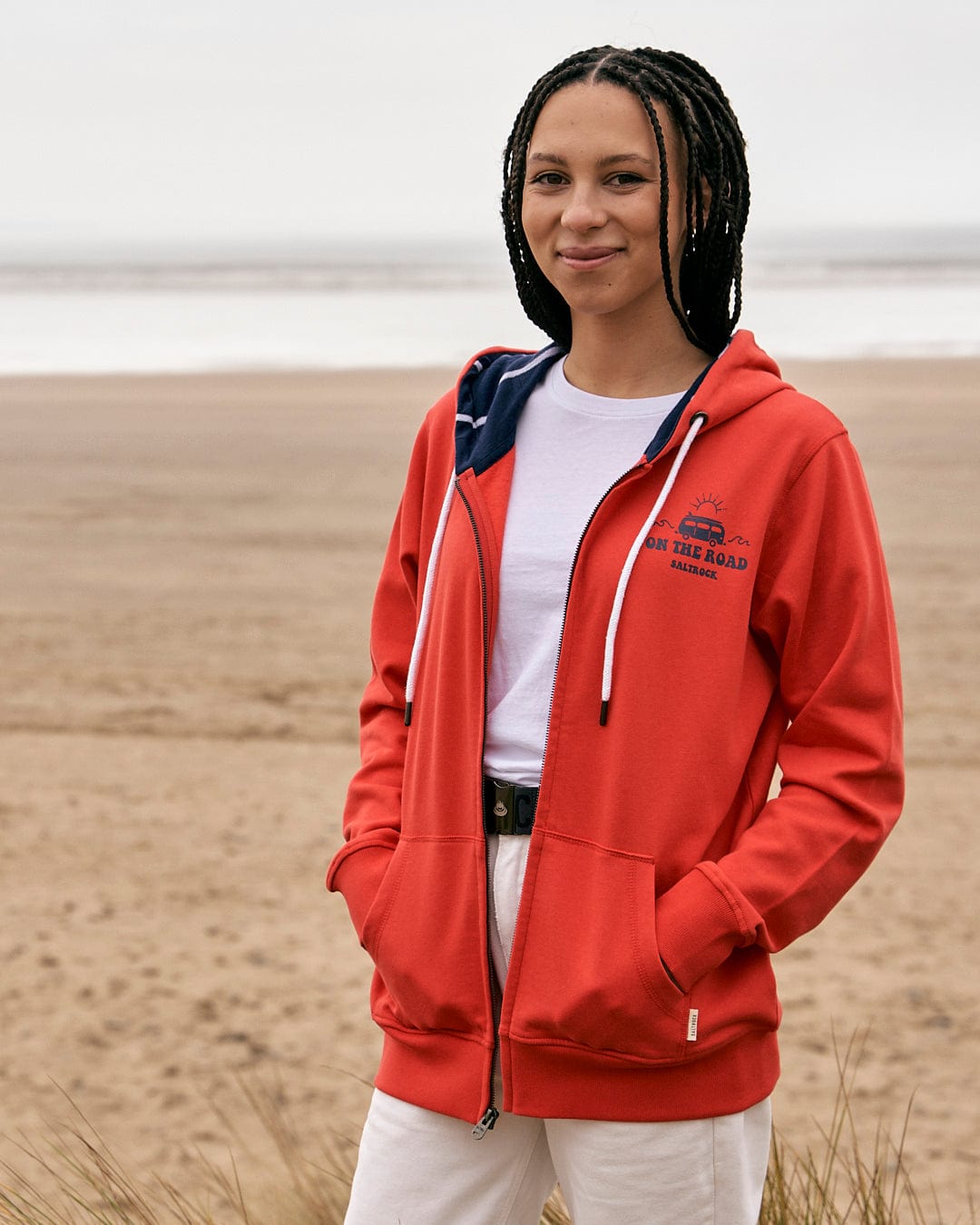A woman wearing a Saltrock On The Road Devon - Womens Zip Hoodie - Red standing on the beach.