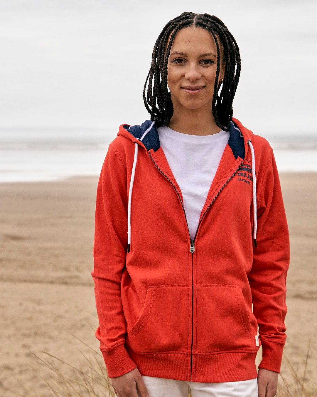 A woman wearing a Saltrock On The Road Wales - Womes Zip Hoodie - Red standing on a beach.