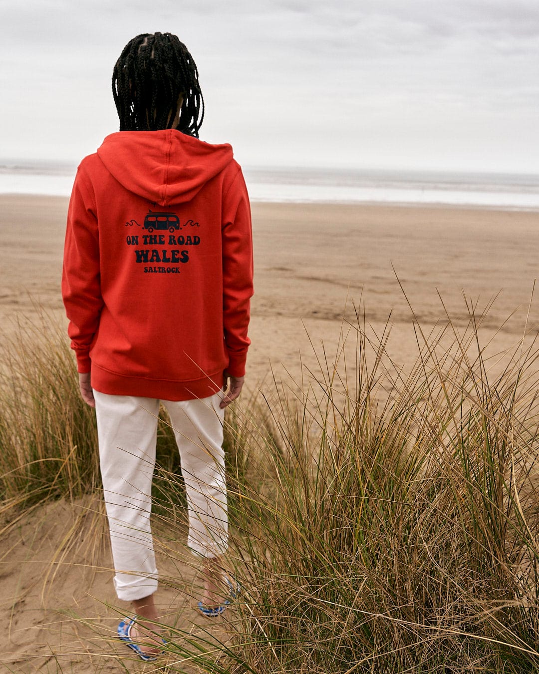 A person standing on a beach wearing a Saltrock On The Road Wales - Women's Zip Hoodie - Red.