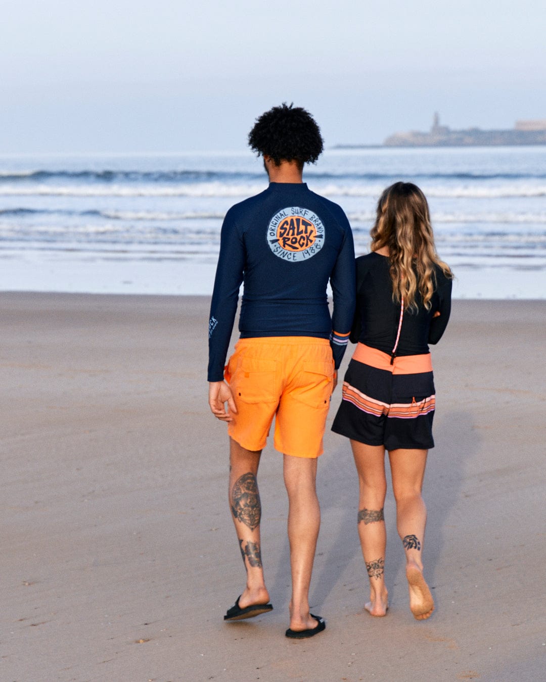Two people in casual beachwear, including SR Original - Recycled Mens Long Sleeve Rashvest - Blue boardshorts by Saltrock, walking on the sand towards the ocean, with a lighthouse visible in the distance.