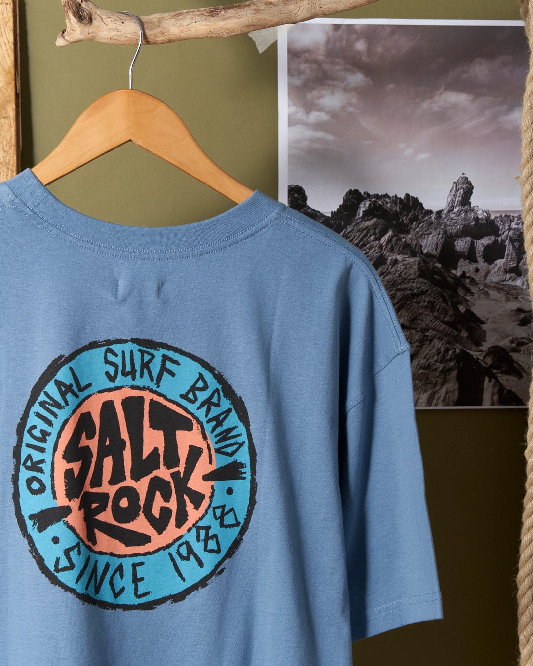 A **SR Original - Mens Short Sleeve T-Shirt - Blue** with the "Saltrock" logo, in an oversized fit, hangs on a wooden hanger in front of a mountainous landscape photograph.
