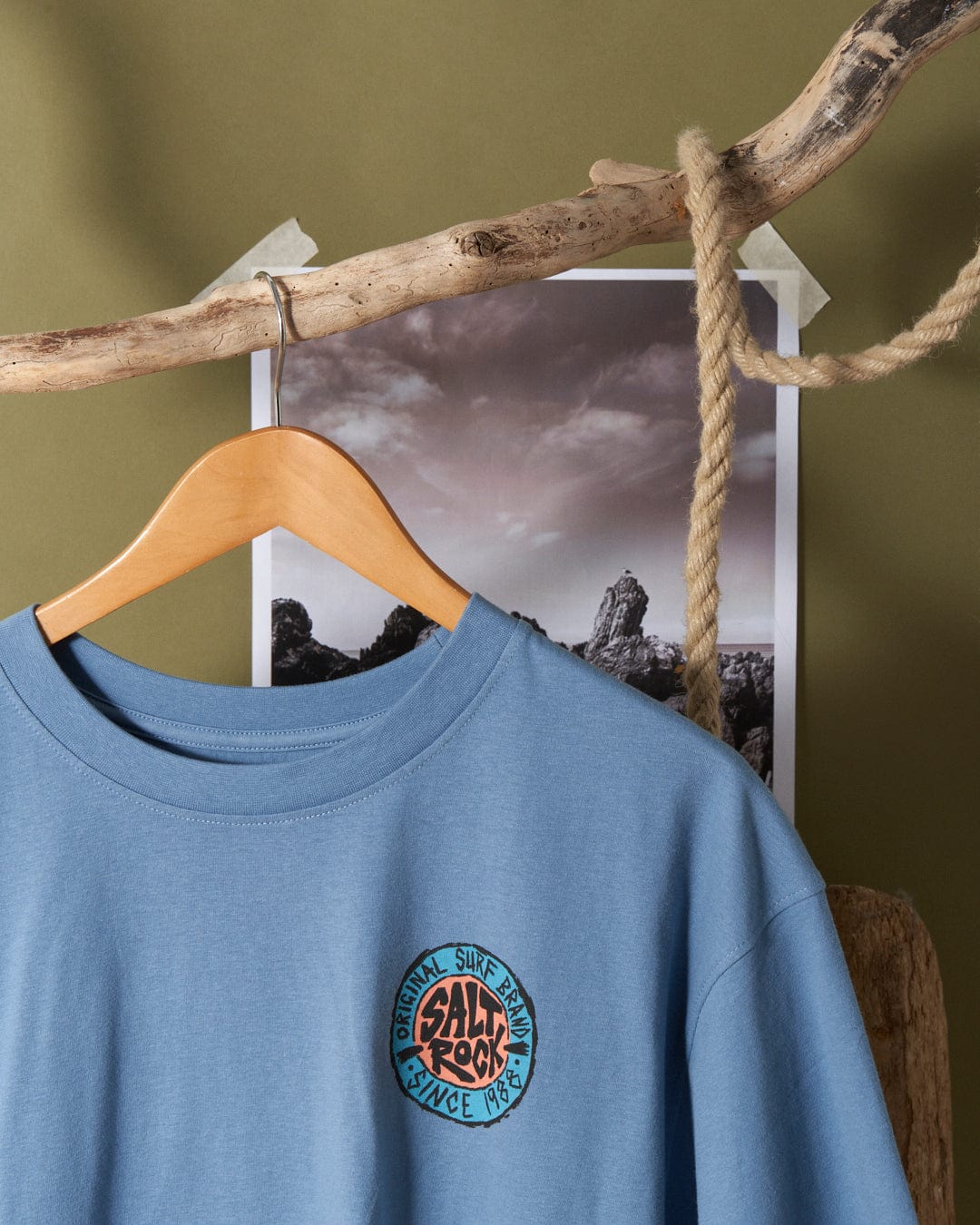 Saltrock SR Original - Mens Short Sleeve T-Shirt - Blue with logo hanging on a wooden hanger against a backdrop with a landscape image.