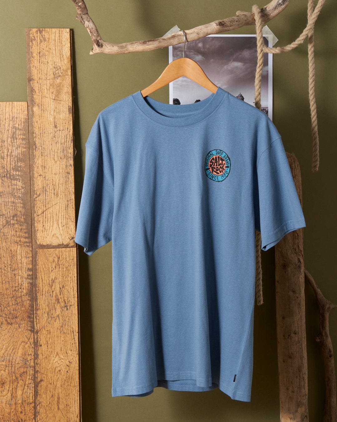Blue Saltrock SR Original Mens Short Sleeve T-Shirt with a logo on the chest, displayed on a hanger against a wooden and green background.