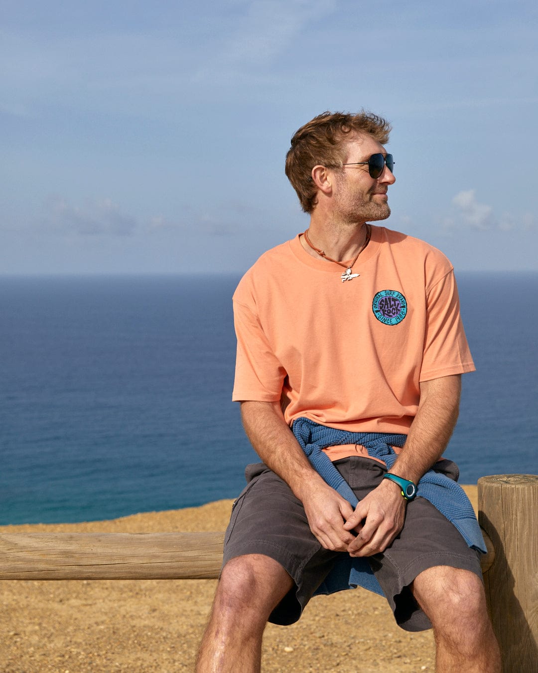 Man sitting on a wooden railing by the sea, enjoying the view on a sunny day, wearing an oversized Saltrock SR Originals mens short sleeve t-shirt in peach with a retro surf graphic.