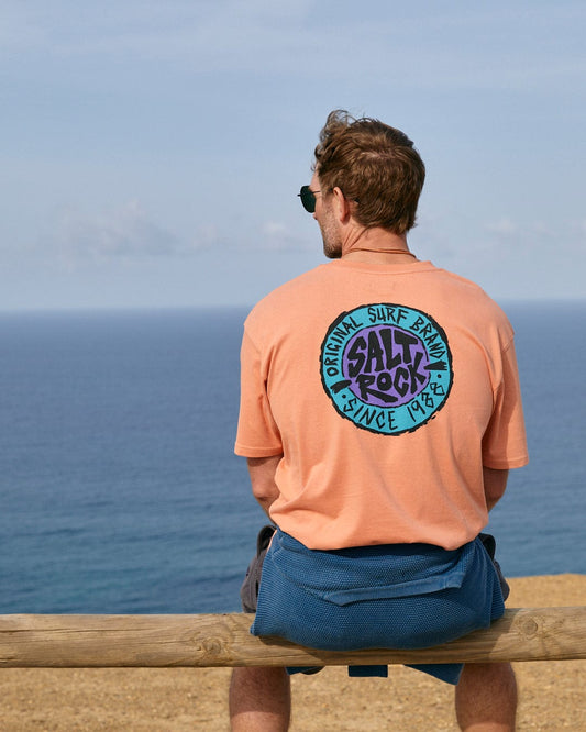 Man sitting on a wooden railing overlooking the sea while wearing an oversized, peach-colored SR Originals - Mens Short Sleeve T-Shirt with a retro surf graphic from Saltrock and sunglasses.