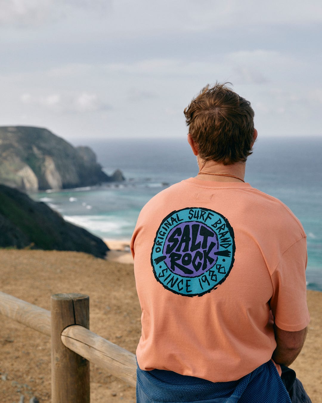 Person in a SR Originals - Mens Short Sleeve T-Shirt - Peach with a retro surf graphic Saltrock logo looking out at a coastal landscape.