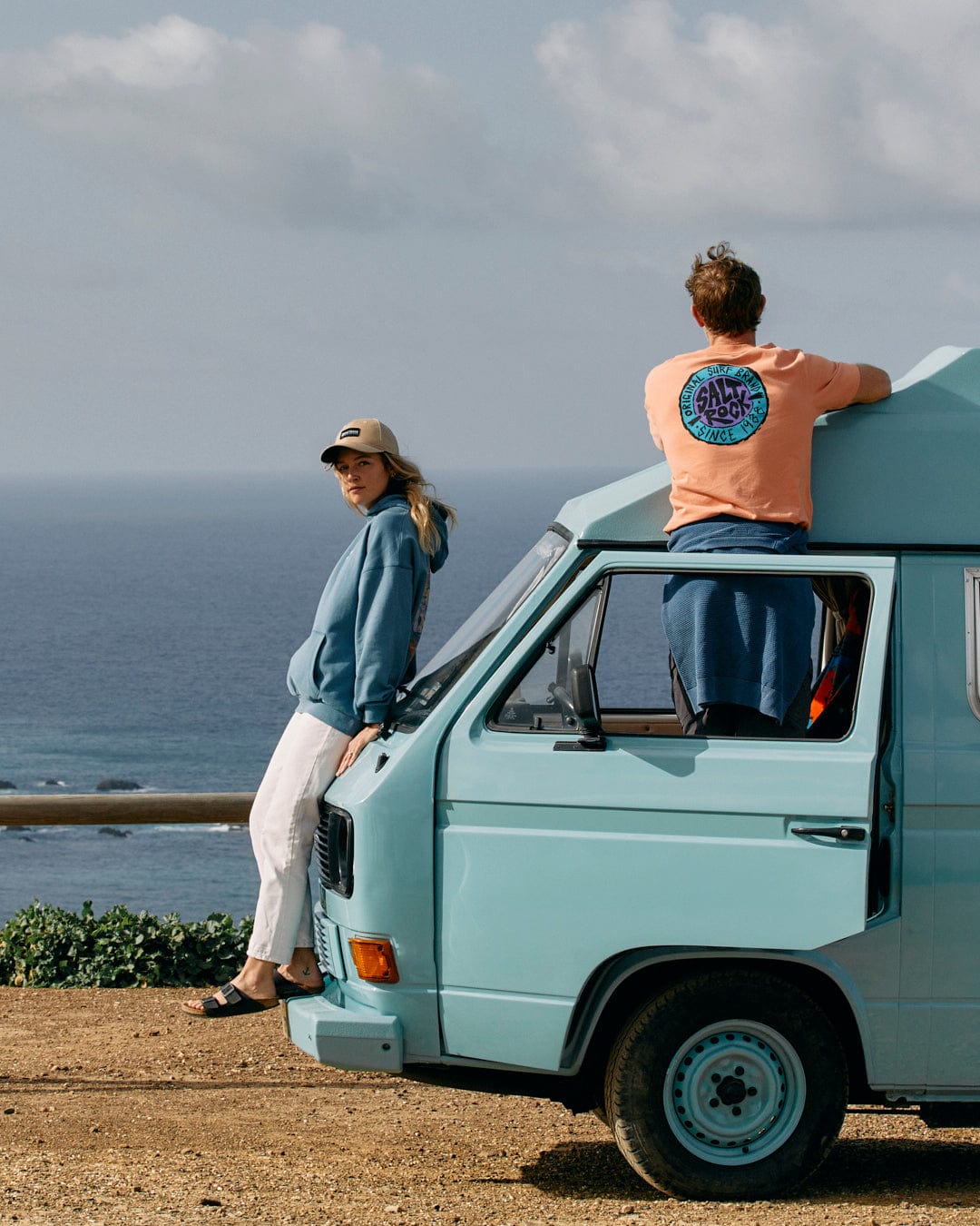 Two people leaning on an oversized blue van featuring a retro surf graphic, enjoying a scenic ocean view wearing Saltrock's SR Originals - Mens Short Sleeve T-Shirt in Peach.
