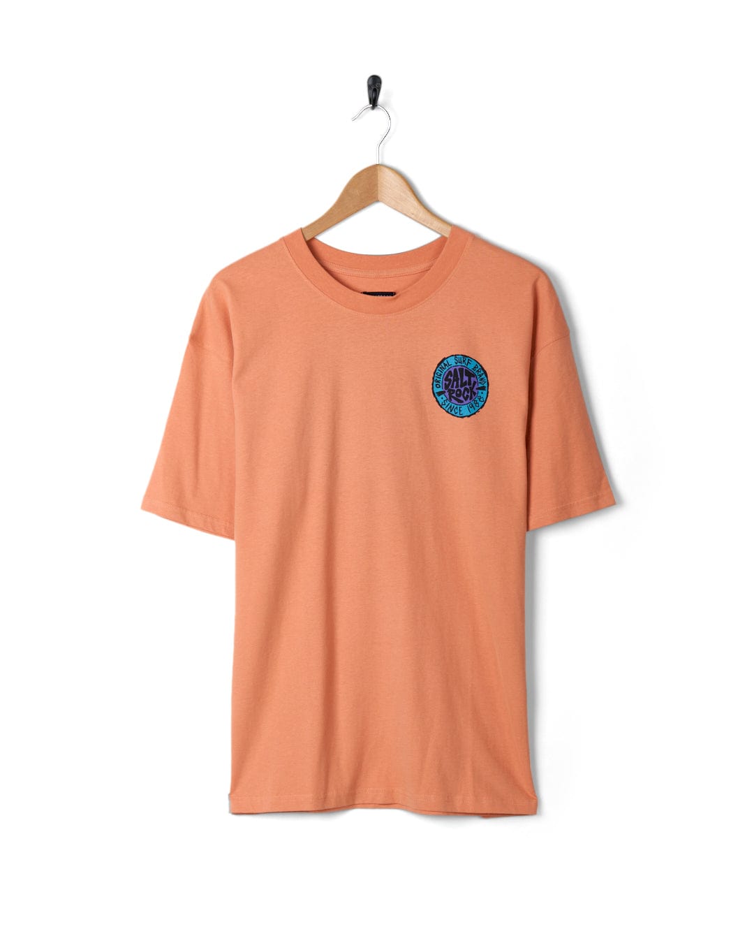 An oversized SR Originals - Mens Short Sleeve T-Shirt in Peach with a small blue emblem on the chest, featuring a retro surf graphic, hanging on a wall-mounted hanger against a white background. Brand: Saltrock