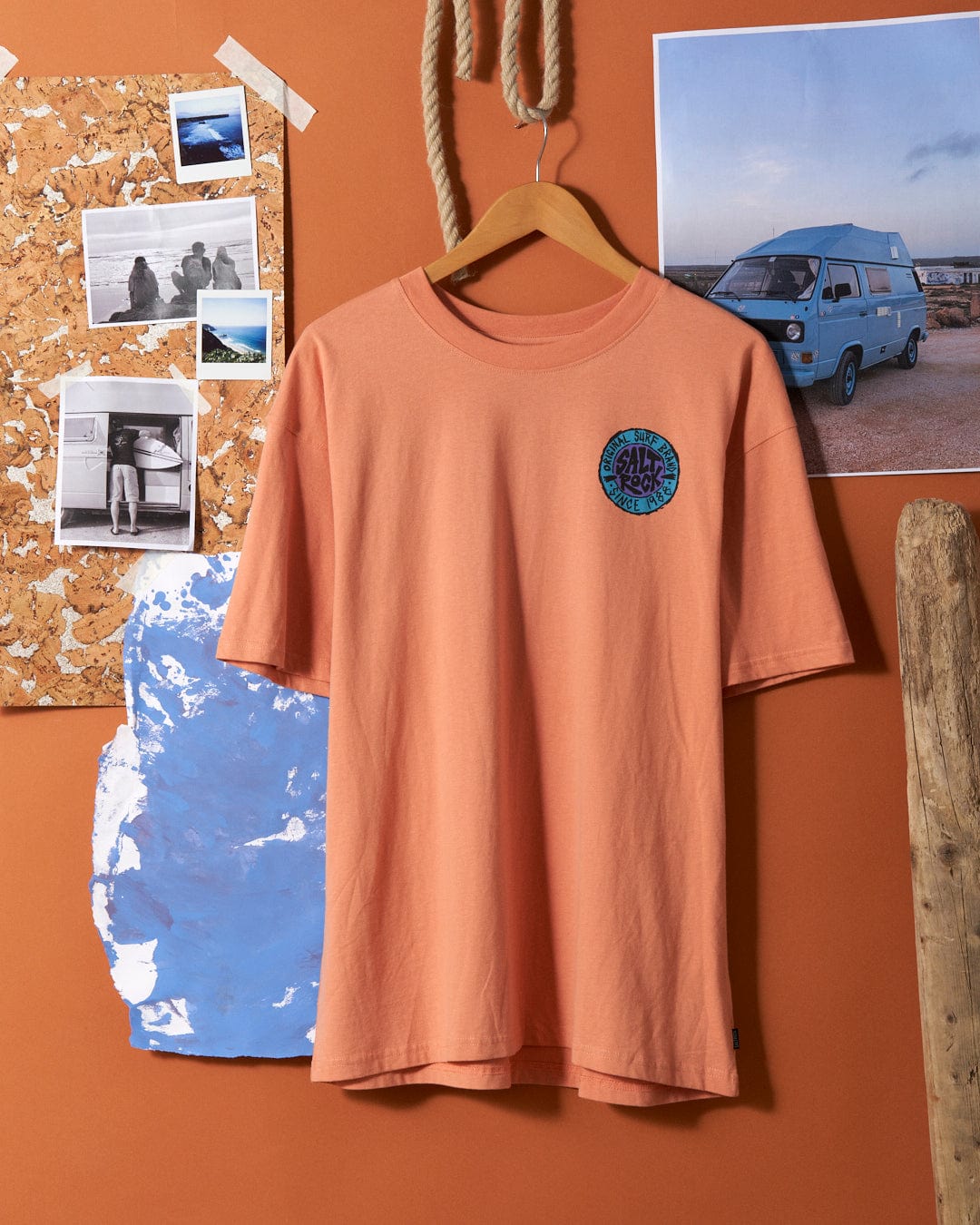 Peach SR Originals oversized t-shirt with a retro surf graphic Saltrock logo on the chest, displayed on a wall beside a corkboard with photos and a blue-painted piece of wood.