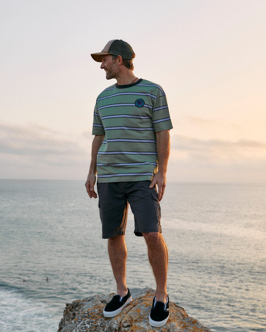 Man standing on a rocky outcrop overlooking the sea at sunset, wearing an Saltrock SR Original - Mens Short Sleeve T-Shirt in Green with a retro surf badge.