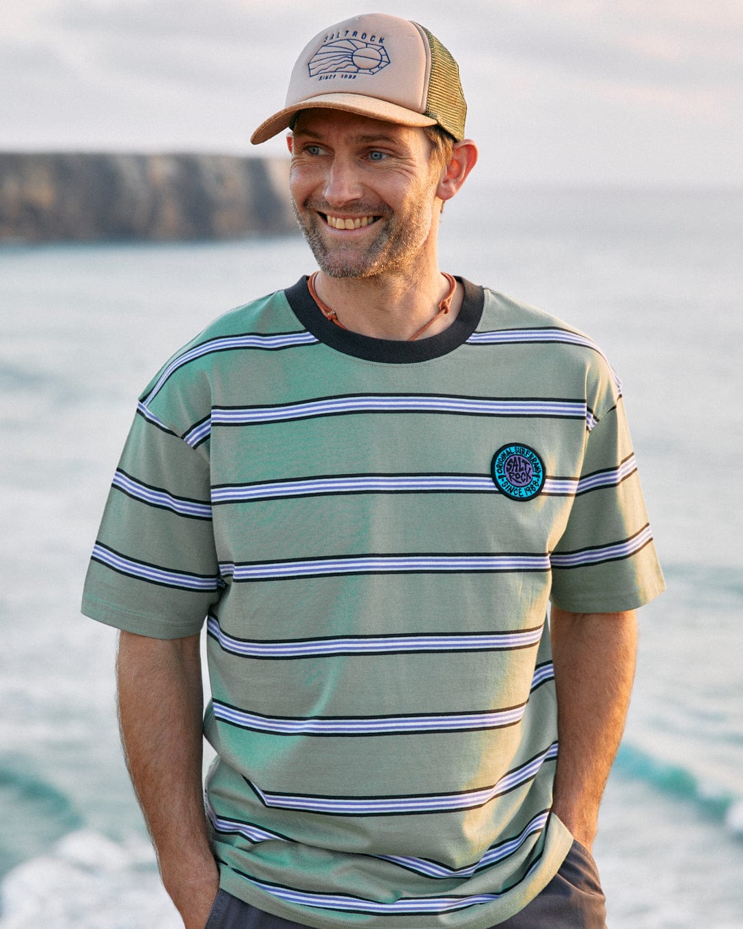 Man in a striped shirt with Saltrock's SR Original - Mens Short Sleeve T-Shirt - Green and cap smiling by the sea.