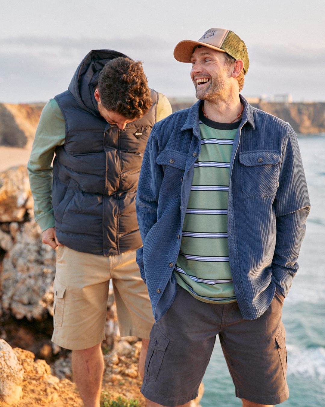 Two men standing on a cliffside, one smiling and looking to the side, the other looking down and adjusting his oversized fit Saltrock Original - Mens Short Sleeve T-Shirt in Green.