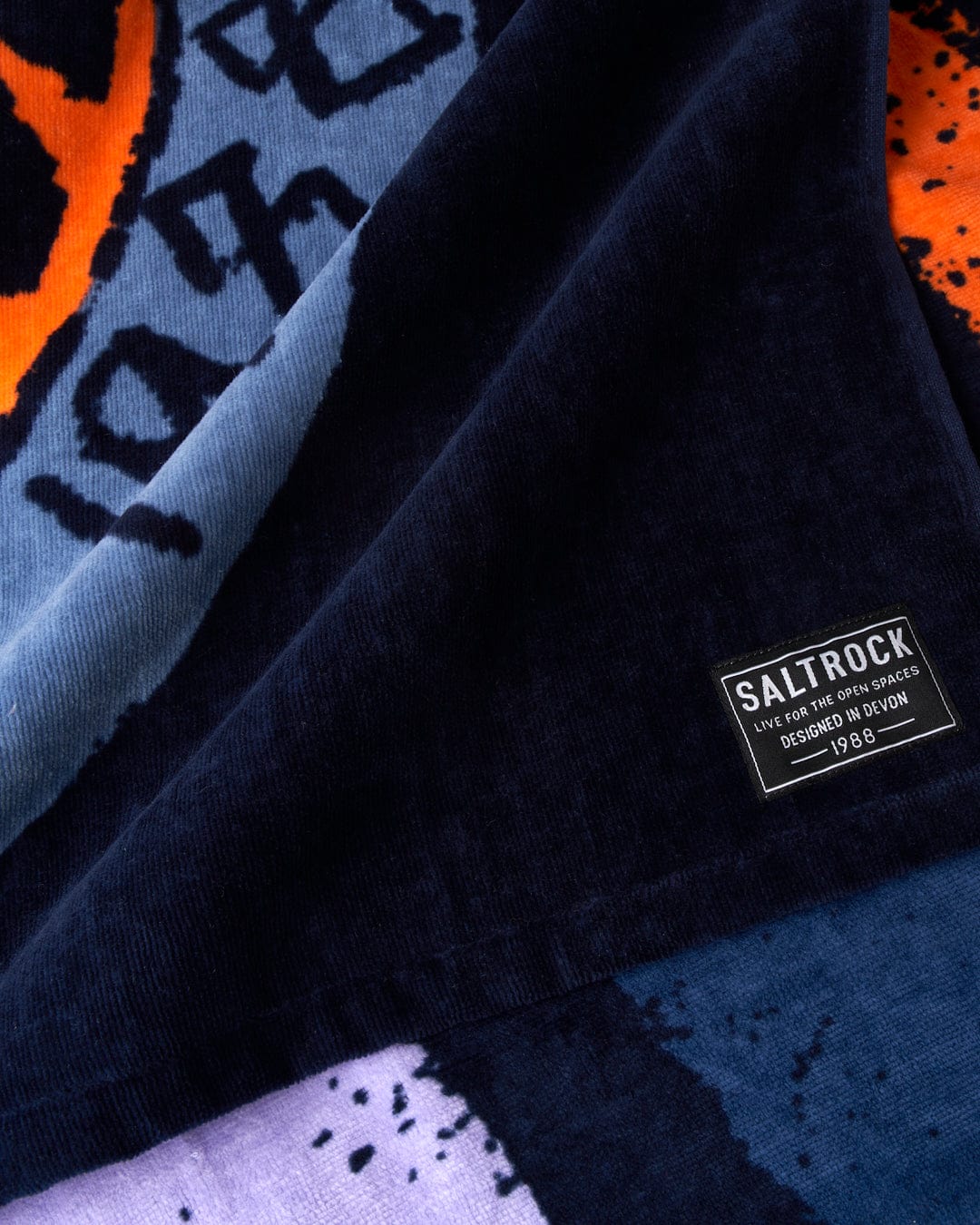 Close-up of a Saltrock SR Original - Towel - Blue clothing label on a dark fabric background with partial view of a soft towelling patterned garment.