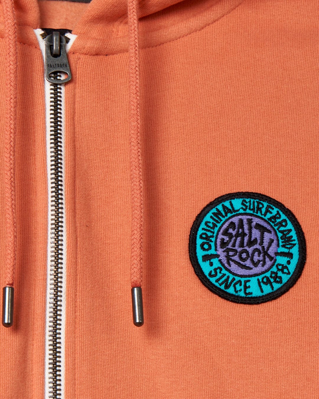Close-up of a coral-colored SR Original - Recycled Mens Zip hoodie - Orange with a zipper and a circular "Saltrock retro surf brand" patch on the left side.