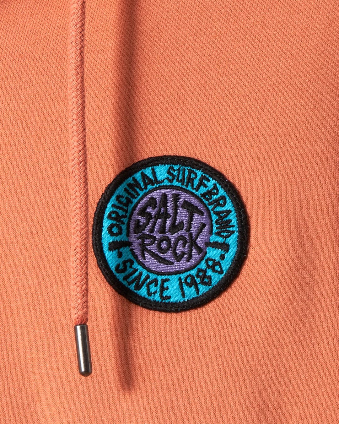 Close-up of a black circular patch reading "Saltrock original Saltrock retro surf since 1989" stitched onto an orange SR Original - Recycled Mens Zip hoodie, with a drawstring visible.