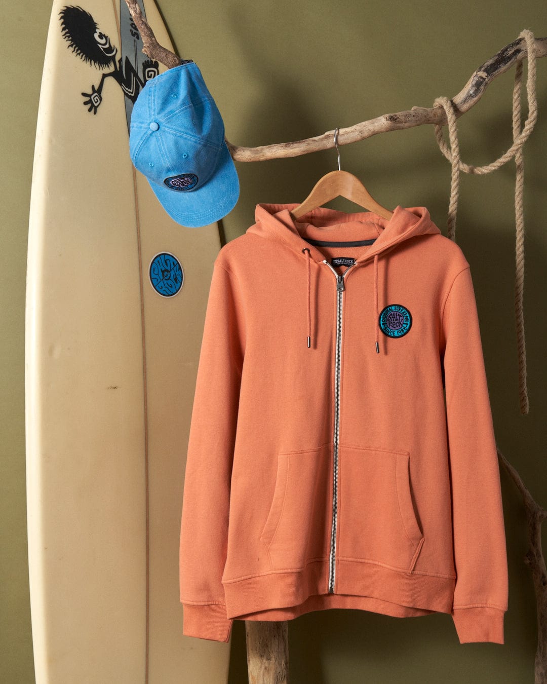 Saltrock orange hoodie with branded metal eyelets and blue cap hanging on a branch next to a Saltrock retro surfboard against a green wall.