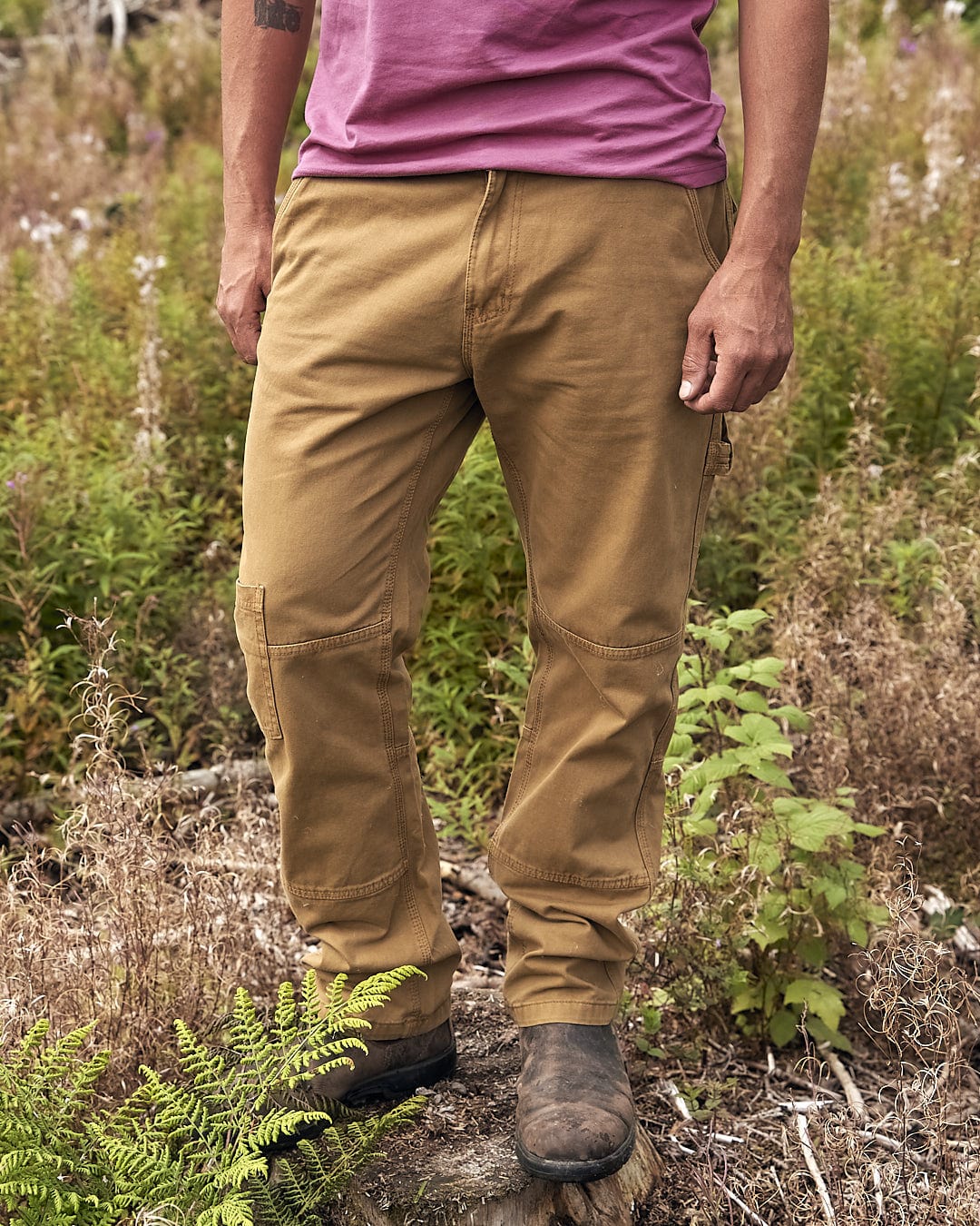 A man standing in a field wearing Saltrock's Oslo - Mens Carpenter Trouser - Brown with detailed stitching.