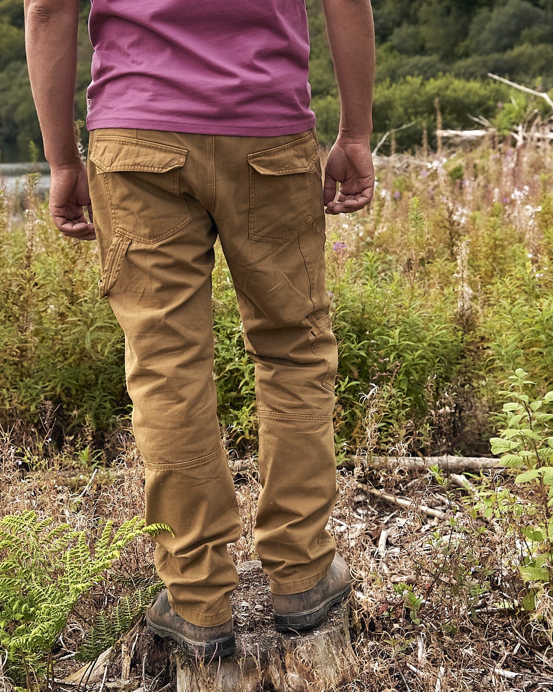 A man wearing Saltrock Oslo - Mens Carpenter Trouser - Brown pants standing on top of a tree stump.