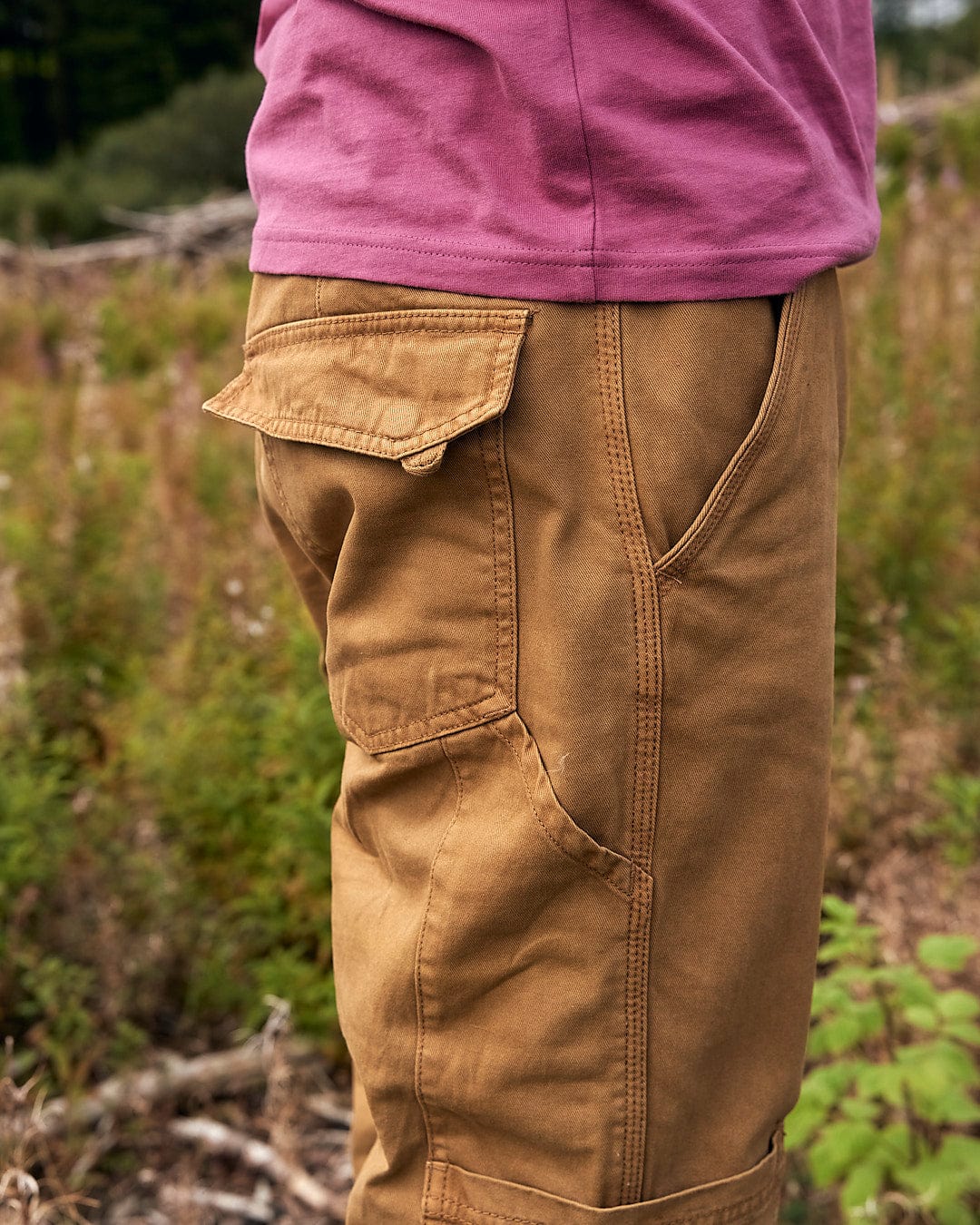 A man standing in the woods wearing a pair of Saltrock Oslo - Mens Carpenter Trouser - Brown with detailed stitching.