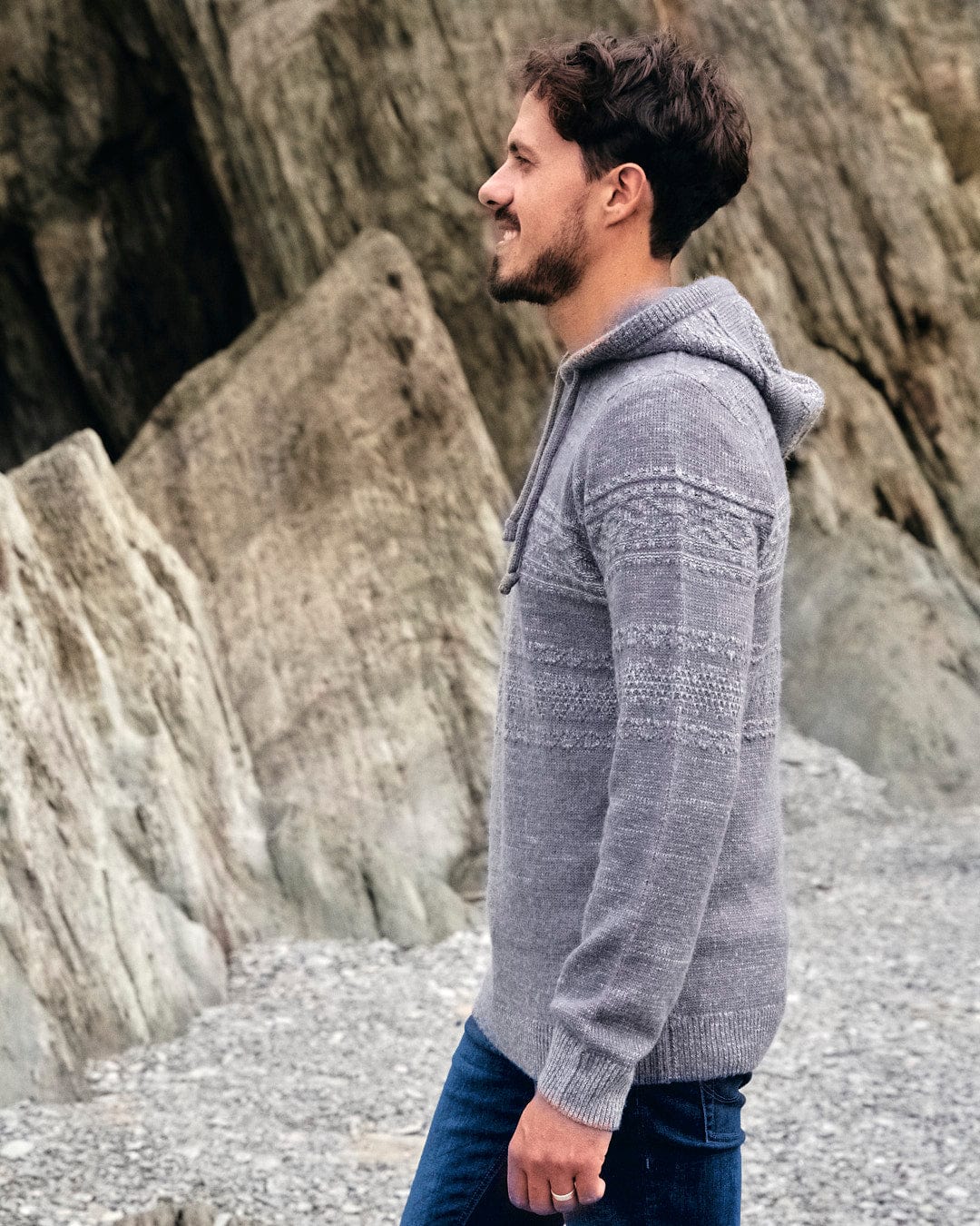 A man wearing a Saltrock - Paxton - Mens Knitted Hoodie - Grey, standing on a rocky beach.
