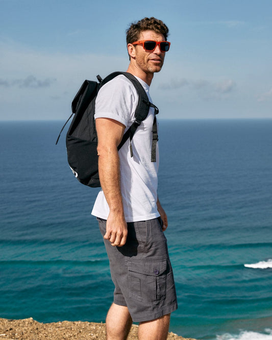Man with Saltrock Penwith II - Mens Cargo Shorts - Dark Grey backpack standing on a coastal cliff overlooking the ocean.