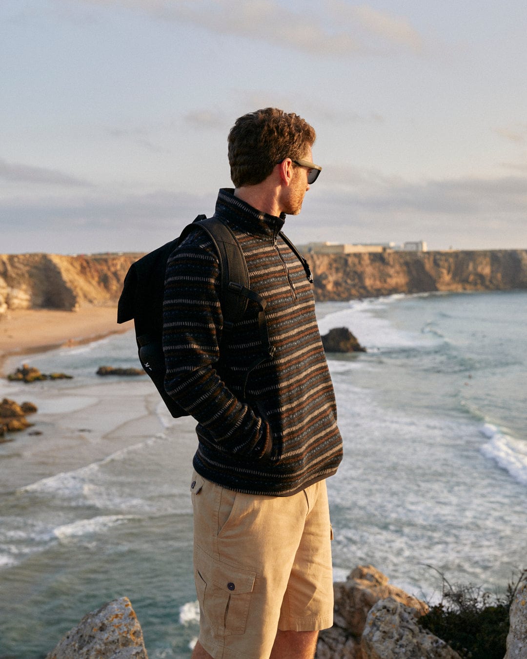 Man with Penwith II - Mens Cargo Shorts - Cream by Saltrock, standing on a cliff overlooking the sea at sunset.