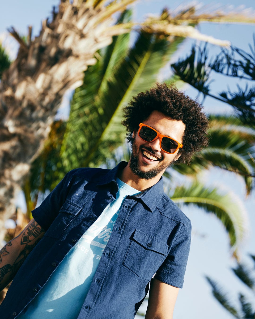 A young man with an afro wearing sunglasses and a Saltrock Polperro - Mens Short Sleeve Shirt - Blue smiles in a sunny setting with a palm tree in the background.