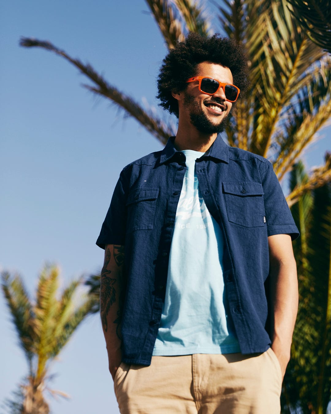 A man with curly hair wearing sunglasses, a Polperro - Mens Short Sleeve Shirt - Blue with a Saltrock woven label over a t-shirt, and beige shorts, is standing outdoors with palm trees in the background.