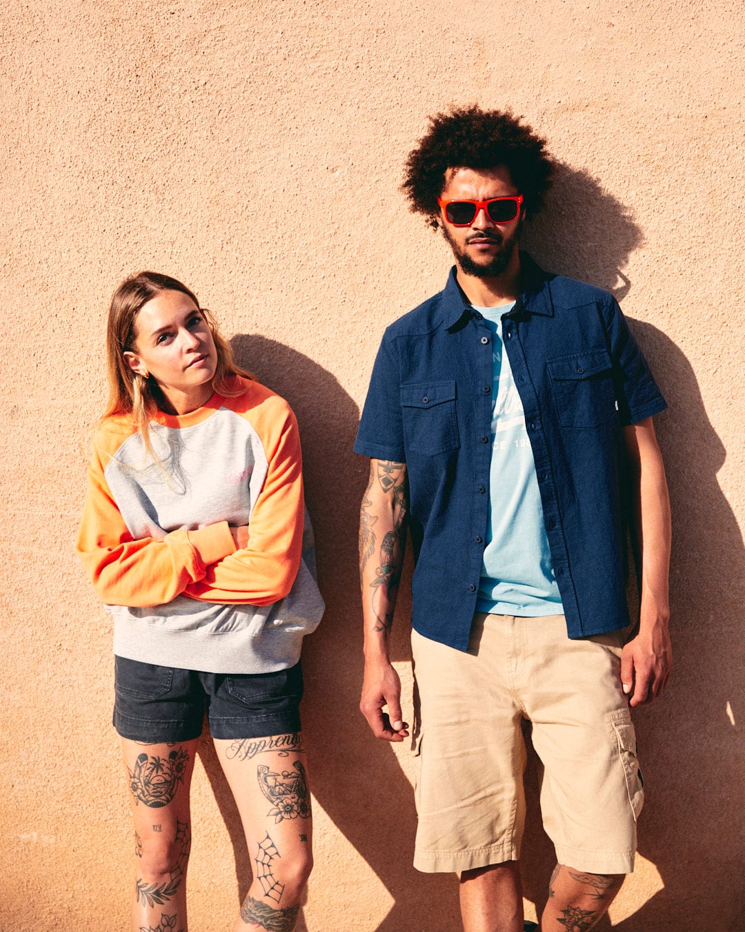 Two young adults, one with tattoos and one with an afro, standing against a sunlit orange wall, dressed in casual summer clothing made of waffle textured fabric including the Saltrock Polperro Mens Short Sleeve Shirt in Blue.
