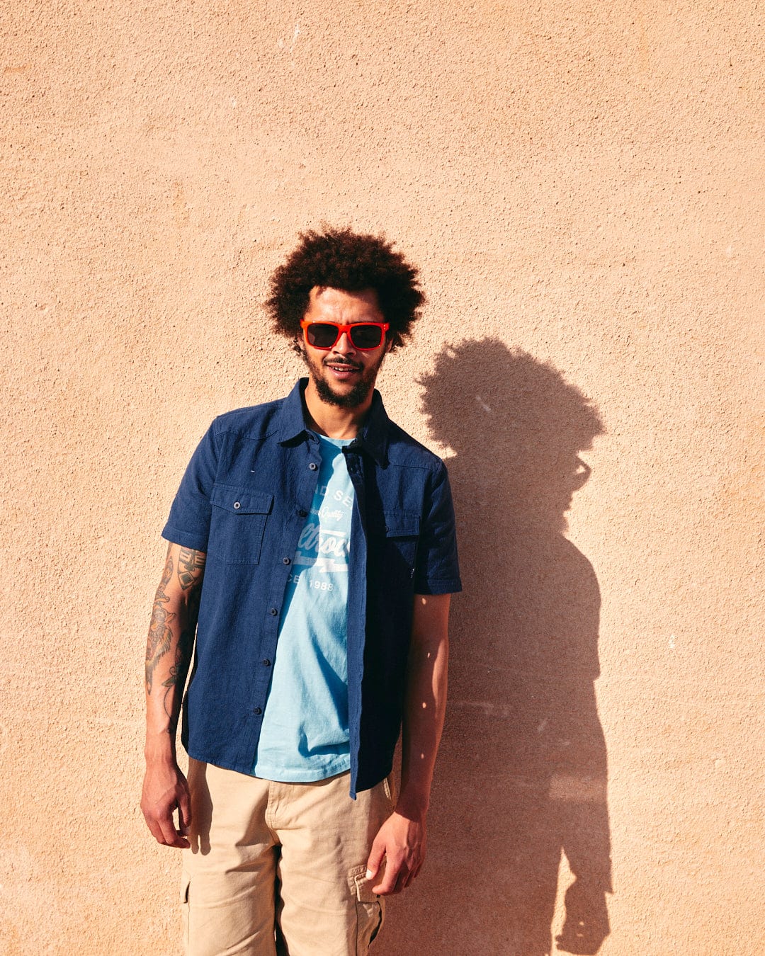 A man with curly hair wearing sunglasses and a Saltrock Polperro - Mens Short Sleeve Shirt in Blue stands against a light-colored wall, casting a shadow.