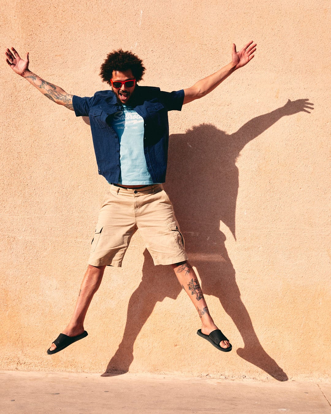 A joyful man with sunglasses and curly hair jumps mid-air against a beige wall, casting a dynamic shadow on the Saltrock Polperro - Mens Short Sleeve Shirt in Blue.