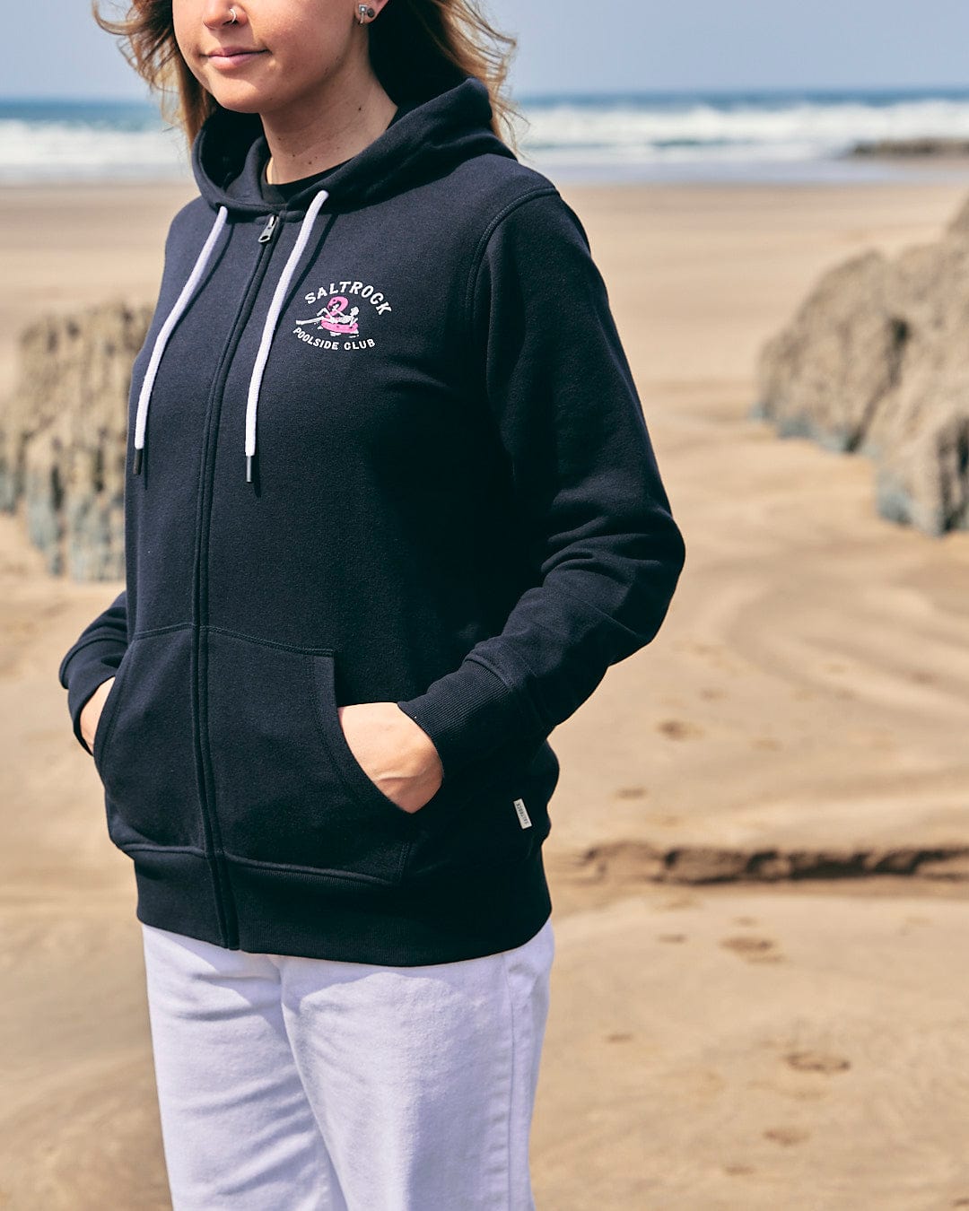 A woman wearing a Lee-Ann - Womens Zip Hoodie - Black by Saltrock standing on the beach.