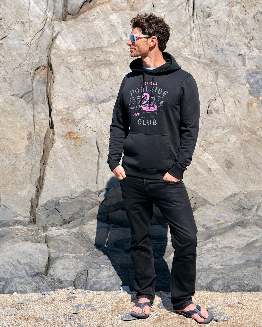 A man wearing a Saltrock Poolside - Mens Pop Hoodie - Black standing next to a rock.