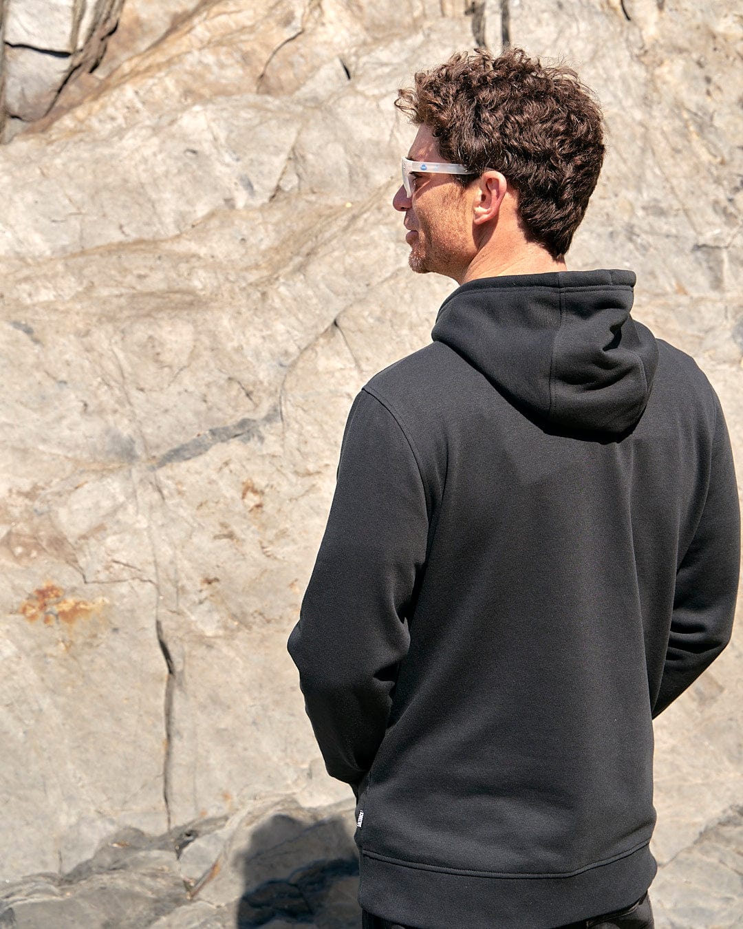 A man wearing a Saltrock Poolside - Mens Pop Hoodie - Black standing next to a rock.