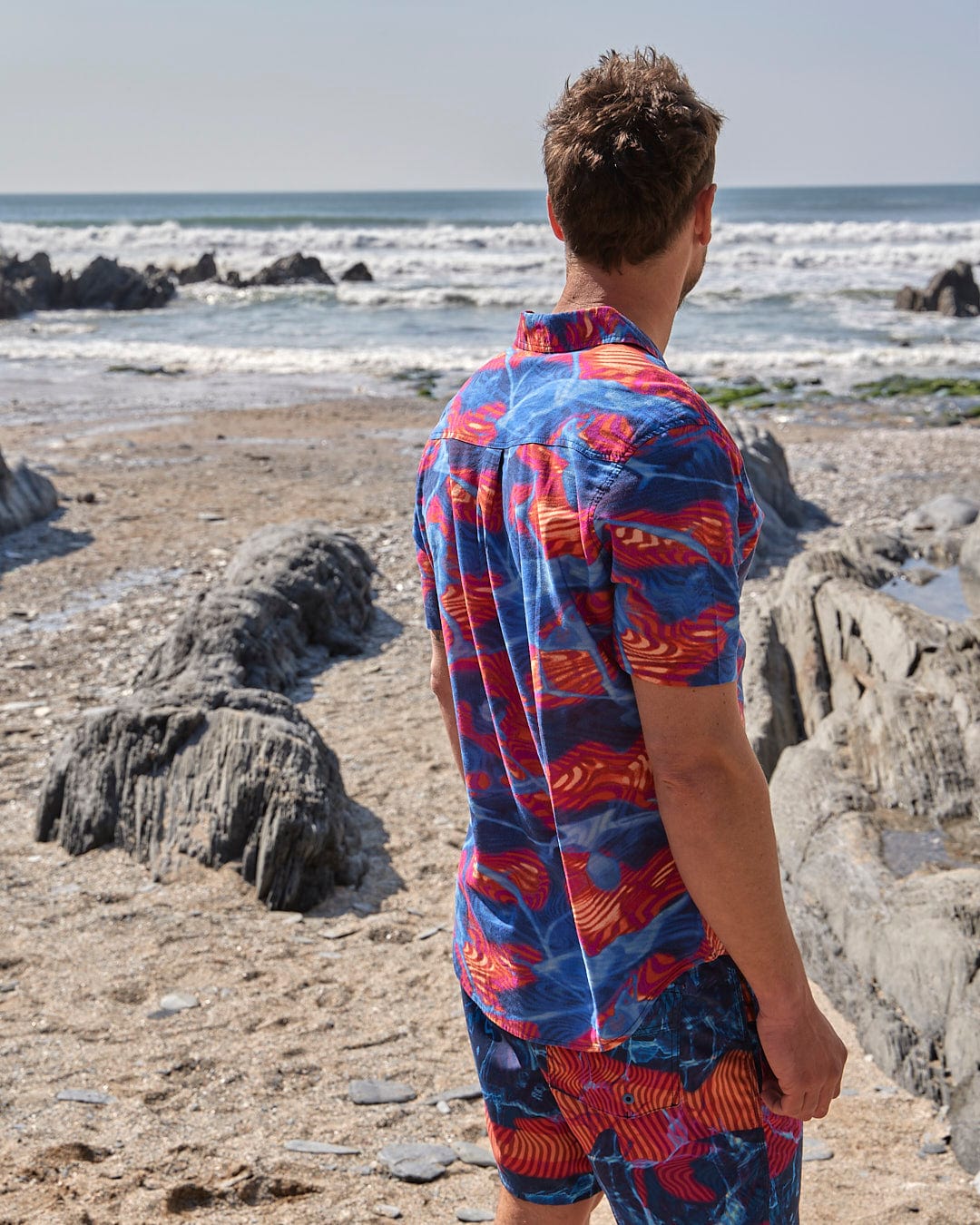A man standing on a beach wearing a Saltrock Poolside - Mens Short Sleeve Shirt, a bright and fun style, made of lightweight fabric.