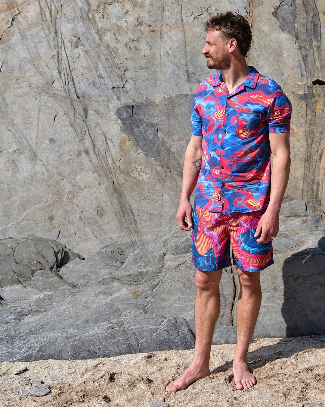 A man standing on a beach wearing a Saltrock Poolside - Mens Short Sleeve Shirt and shorts made with lightweight fabric.