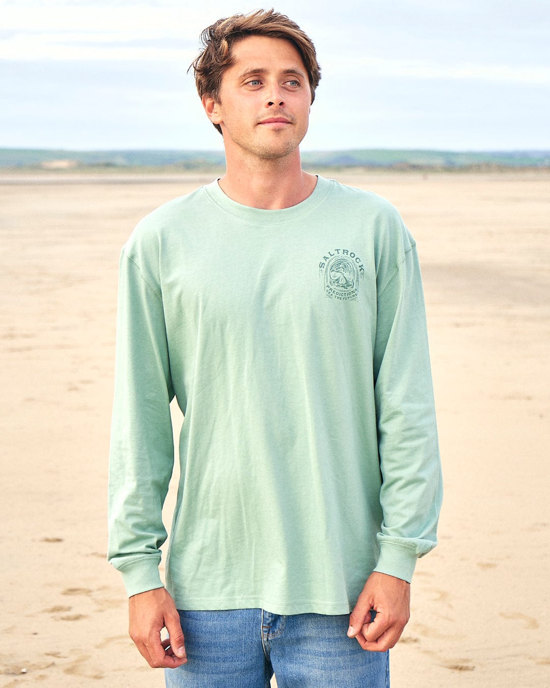 A man wearing a Saltrock Predictions - Mens Long Sleeve T-Shirt - Light Green standing on the beach.